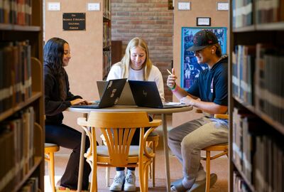 Three students sitting together at a table in the library with their laptop computers open.