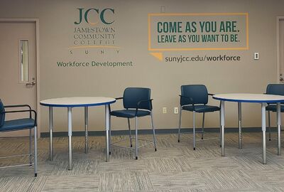 A sitting area with tables and chairs with SUNY JCC Workforce Development logos on the wall in the background.