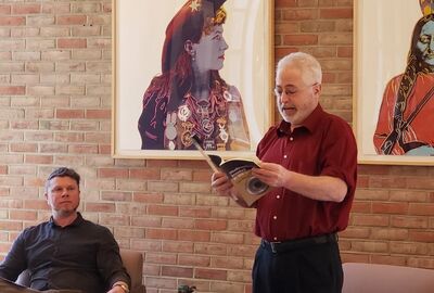 A man reads a book aloud in a library while a seated person looks on.