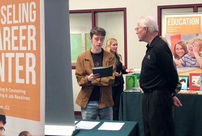 A student writes on a notebook while talking to a faculty member at a college open house event.