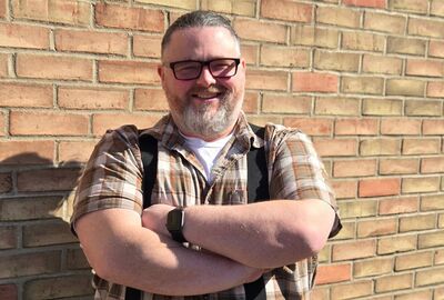 A person smiling with arms crossed with a brick wall in the background.
