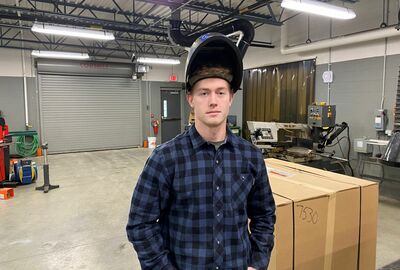 A young man wearing a welding hood stands above his face inside a manufacturing area, surrounded by shelves and industrial equipment.