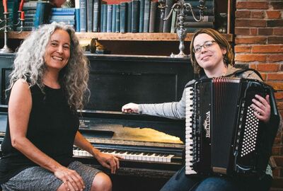 Two people sit at near a piano with one holding an accordion.