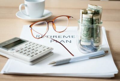 A stack of papers, a calculator, pen, glass cup filled with folded dollar bills, eye glasses and a coffee cup and foster arranged on a wood table.