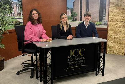 Three students sit at a desk on a college TV studio set with a SUNY JCC logo on the front of the table.