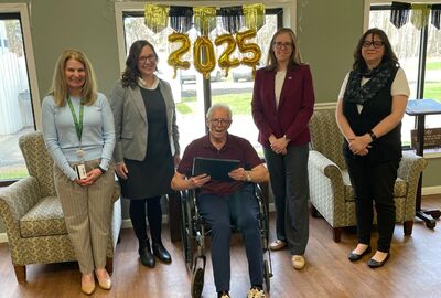 Four women stand by a man in a wheelchair holding a college diploma  with a 2025 balloon hanging in the background.