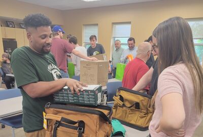 A person takes items out of a tool bag while another watches.