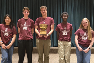Six classmates wearing the same colored maroon t-shirts pose with one in the middle holding a trophy.