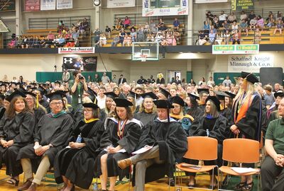 College employees and students dressed in graduation caps and gowns sit in rows of cheers in a gymnasium during commencement.