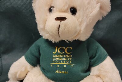 A small stuffed bear wearing a green and gold SUNY JCC alumni shirt.