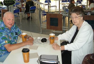 Two older adults sit at a table in a café, each with a coffee cup, looking toward the camera. The background shows other people seated at tables.