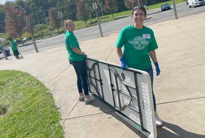 Two smiling volunteers wearing green “Remote Area Medical” t-shirts and blue gloves carry a folding table outside on a sunny day, preparing for a community clinic event.