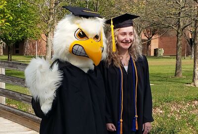 A student wearing a graduation cap and gown poses with JCC's Jayhawk mascot, also wearing a graduation cap and gown, on campus.