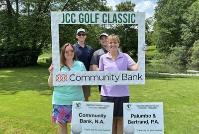 Four smiling golfers pose together outdoors on a sunny day, holding a large photo frame that reads "JCC Golf Classic" with the Community Bank logo below. Two sponsor signs in front of them read "Community Bank, N.A." and "Palumbo & Bertrand, P.A.," thanking them for their support of the Jamestown Community College Classic Golf Tournament. Lush green trees and a small pond are visible in the background.