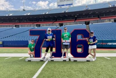 Four people pose on a football field in front of large block numbers “716,” which represent the Western New York area code. The group is standing at Highmark Stadium, home of the Buffalo Bills, wearing Bills gear and green SUNY JCC shirts. The stands are mostly empty under a blue sky with scattered clouds.   