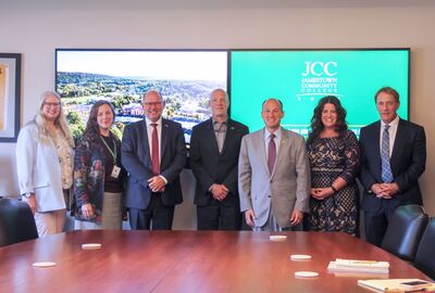 Group photo of seven people, including the Sweden U.S. ambassador, standing in a conference room at SUNY Jamestown Community College. Two large digital screens behind them display a scenic aerial view and the JCC logo. An American flag is visible on the right.