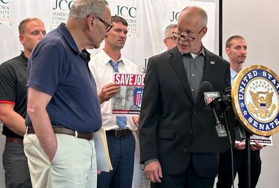 Two men together at a podium and microphone with others in the background holding signs supporting Job Corps.