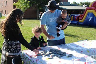 A family gathers around a table covered with a floral tablecloth at an outdoor event. A woman and a young child pick up smooth stones from the table, while a man wearing a black hat and light blue "Top Air" shirt holds a baby. In the background, there is a colorful inflatable play structure and a grassy area with trees and buildings.
