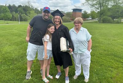 A group of four people stands smiling on a green lawn during a graduation celebration. At the center is a woman wearing a black graduation cap and gown, holding a diploma and gift bag. A young girl, a man in a Buffalo Bills cap, and an older woman in a light-striped shirt stand beside her, all posing happily. Trees and a brick building are visible in the background under a partly cloudy sky.