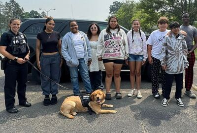 A group of police officers, a therapy dog, and smiling teenagers stand in front of a vehicle in a parking lot. 