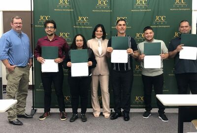 Seven individuals stand in a row, five holding certificates, against a green JCC Workforce Development backdrop, conveying pride and achievement.