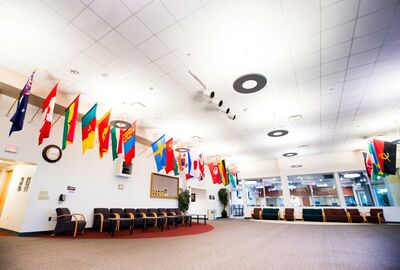A spacious lobby with rows of chairs and numerous international flags hanging from the ceiling. Large windows allow natural light into the room.