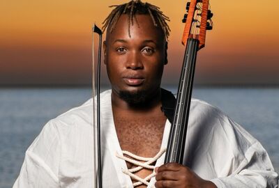 A person with a calm expression holds a string instrument and bow, standing before a tranquil sea at sunset. Wears a white, lace-up shirt. 