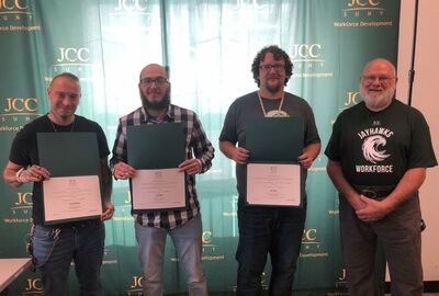 Four men stand smiling in front of a green backdrop with "SUNY JCC Workforce Development" logos. Three hold certificates, celebrating an achievement.