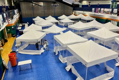 A large indoor space with multiple white tents set up on a blue tarp, resembling a makeshift health clinic. People are working inside a tent.