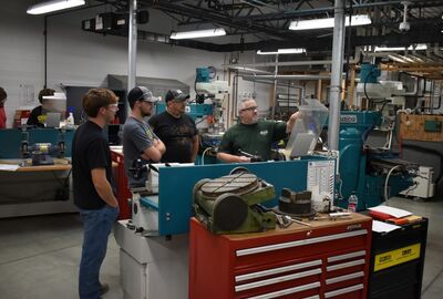 A group of four people stand around a machine in a workshop, as one gestures towards a monitor, indicating a technical discussion. 