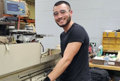A person wearing safety glasses and gloves operates a machine in a workshop. They are smiling, conveying a positive and confident atmosphere.
