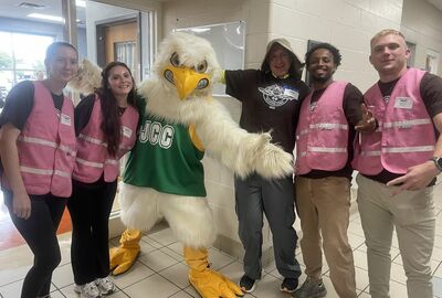 A group of five people and a person in a bird mascot costume pose indoors. Some wear pink vests, smiling, conveying a cheerful and friendly atmosphere.