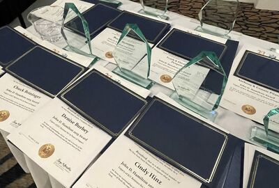 A table displays multiple award plaques and glass trophies neatly arranged. Each plaque is blue with gold writing.