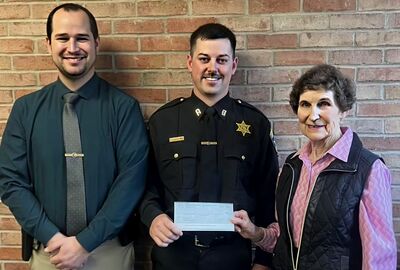 Three people stand smiling against a brick wall. The man in the center, wearing a police uniform, holds a check.