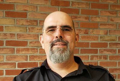 A man with glasses, a grey beard, and a black shirt stands in front of a brick wall, looking at the camera and smiling slightly.