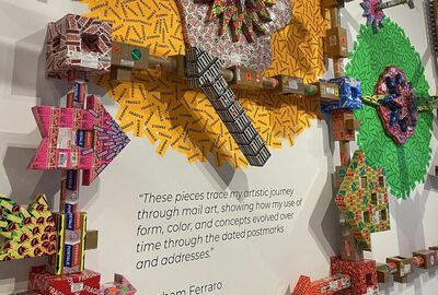A colorful wall sculpture made of various labeled packages and arrows, with an artistic quote by Abraham Ferraro about mail art evolution.