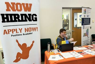 A table with a bright orange tablecloth covered with papers, brochures, and a laptop. A person in colorful clothing sits behind it. A large "Now Hiring" sign stands beside the table, indicating an inviting and energetic job recruitment or career fair atmosphere.