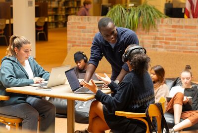 Students collaborate in a library. A man stands, smiling, discussing with a seated woman wearing headphones. Others study quietly nearby. 