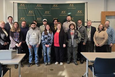 A diverse group of 16 people smiling in front of a green JCC Workforce Development banner. The setting is a graduation ceremony, conveying a positive and professional atmosphere.