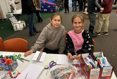 Two smiling young girls sit at a craft table covered with markers, scissors, and washi tape in a busy indoor event. The atmosphere is lively and creative.