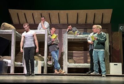 Stage scene with six men in casual clothing on a set resembling military barracks. Some read scripts, others look contemplative,.
