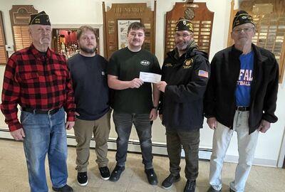 Five men stand indoors, smiling, with two in the center holding a check. Plaques are visible in the background.