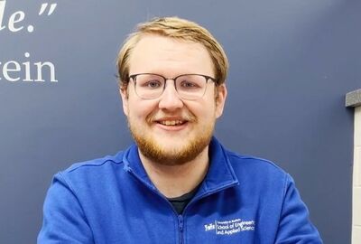 A smiling person with glasses and a beard wears a blue jacket with a logo for the School of Engineering. The background is a blue wall with partial text.