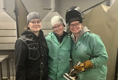 Three women in a workshop wearing safety goggles and gear, including gloves and welding helmets. They are smiling, conveying teamwork and camaraderie.