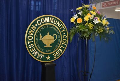 Seal of Jamestown Community College with a lamp design, flanked by vibrant yellow and white flowers on a blue curtain backdrop.