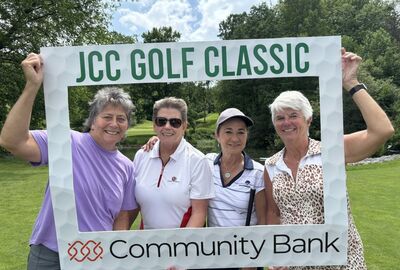 Four smiling women stand together holding a "JCC Golf Classic" frame, with lush green trees in the background, conveying joy and camaraderie.