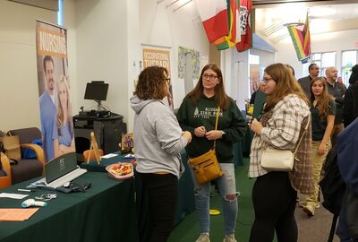 Three people engage at a college open house surrounded by nursing and occupational therapy banners. Tables hold promotional materials. 
