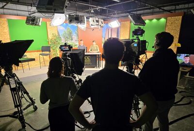 TV studio with two presenters seated at a desk under bright lights. Camera operators and crew stand around, creating a focused atmosphere.