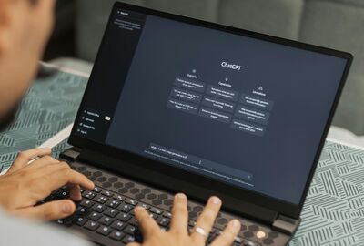 A person types on a laptop displaying the ChatGPT interface. The screen shows features and capabilities, while the background has a patterned tablecloth.