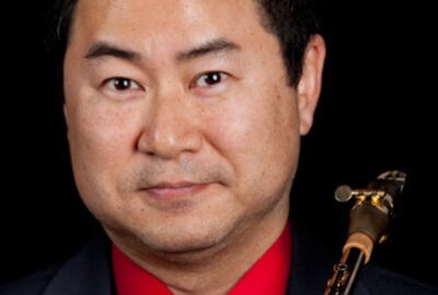 Portrait of a man with a saxophone. He wears a red shirt and dark suit, exuding a calm, focused demeanor. The background is solid black.